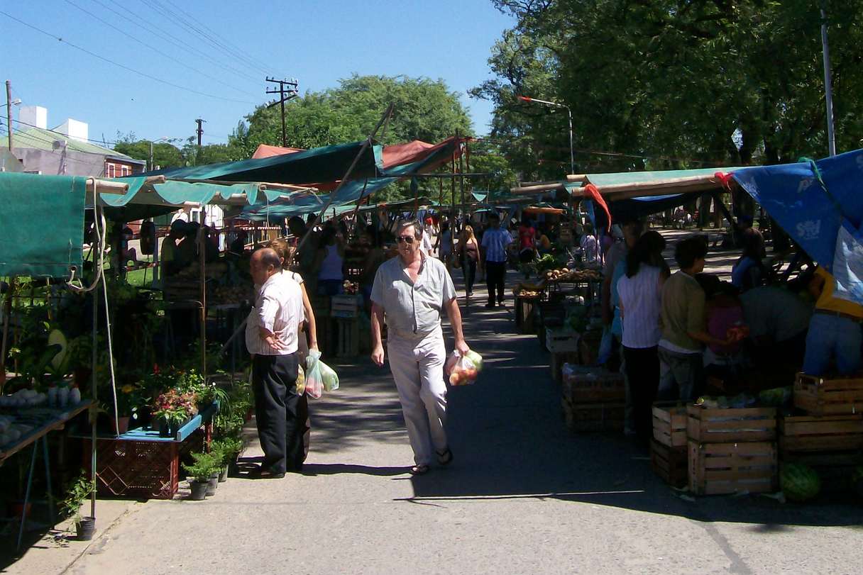 Feria de frutas y verduras al aire libre, bajo toldos de lona, con cajones de madera y gente que camina con bolsas de compras.