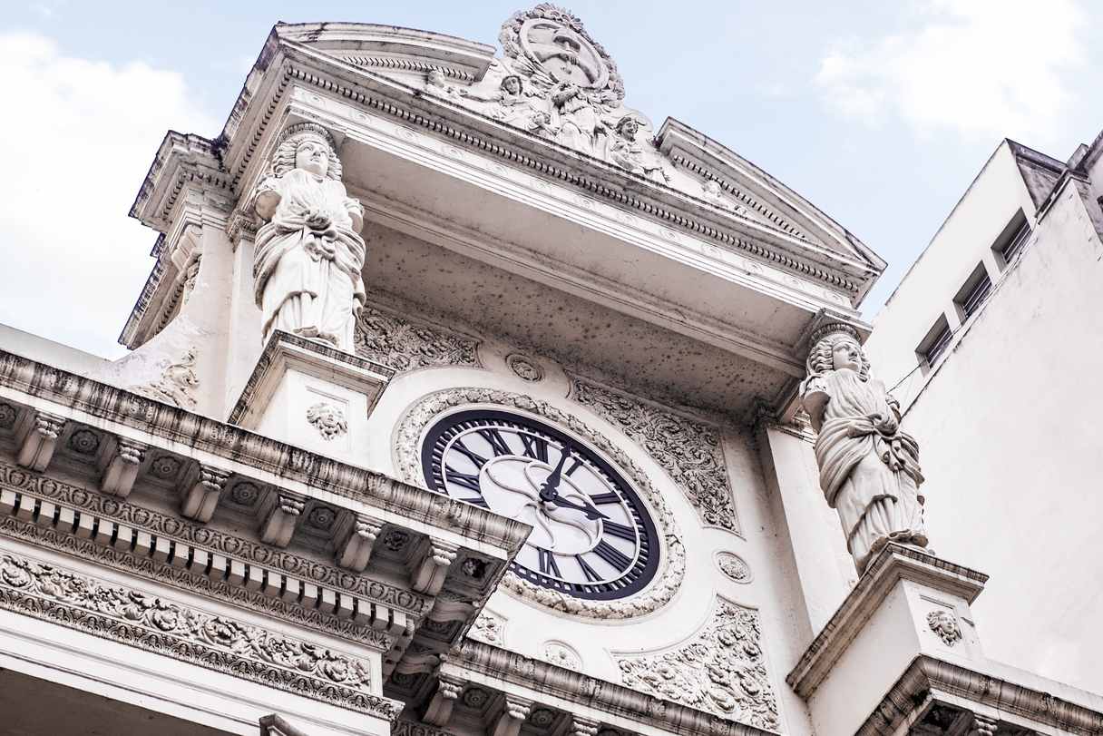 Frente de un edificio visto desde abajo: un reloj de números romanos rodeado de molduras, entre dos figuras femeninas esculpidas que sostienen la cornisa y el frontis.