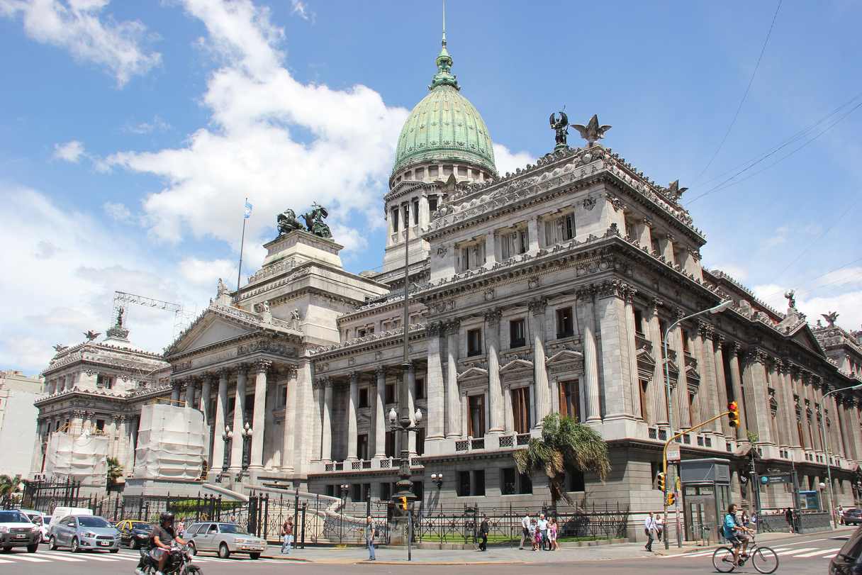Palacio neoclásico de piedra clara con una gran cúpula de cobre verdoso, columnas y grupos escultóricos de bronce sobre los frentes, visto desde una esquina, con autos, un ciclista y peatones en la calle.