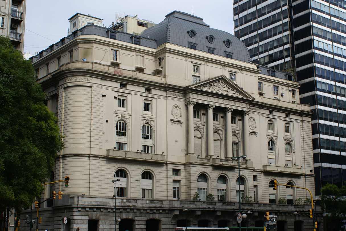 Edificio neoclásico de esquina, con cúpula oscura, frontón esculpido y columnas sobre el ingreso principal; detrás asoma una torre vidriada y en la vereda hay semáforos y árboles.