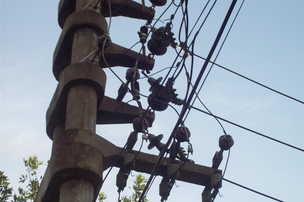 Cabezal de un poste de hormigón visto desde abajo, con ménsulas, aisladores de cerámica y una decena de cables que se cruzan contra el cielo; en el borde inferior asoman las hojas de un árbol.