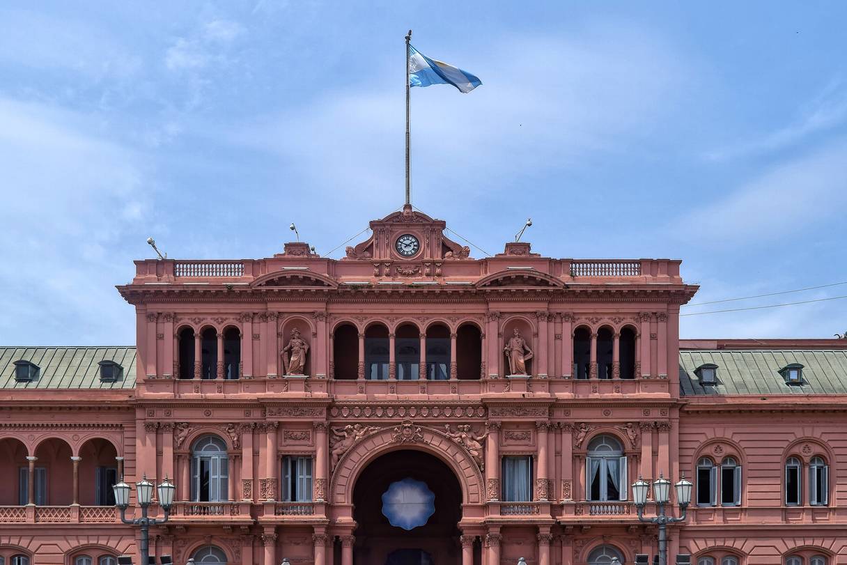 Fachada de un palacio de color rosado con columnas, arcos y balconada: dos estatuas flanquean la galería central, un reloj corona el frontis y una bandera celeste y blanca flamea en el mástil, bajo un cielo azul con cirros finos.