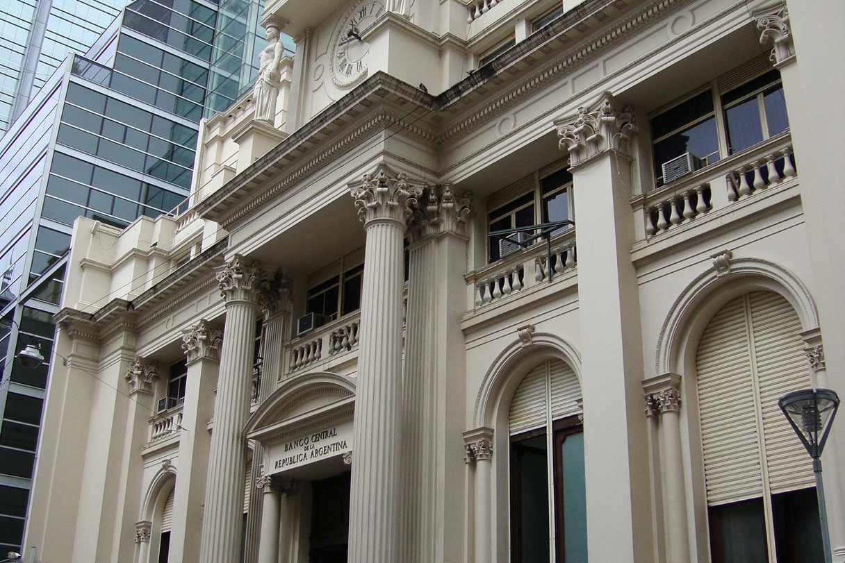 Fachada neoclásica de piedra clara vista desde la vereda, con columnas corintias, balaustradas y ventanas en arco; sobre el acceso, la inscripción tallada «Banco Central de la República Argentina»; arriba, un reloj y una figura esculpida; a la izquierda, un edificio moderno de vidrio.