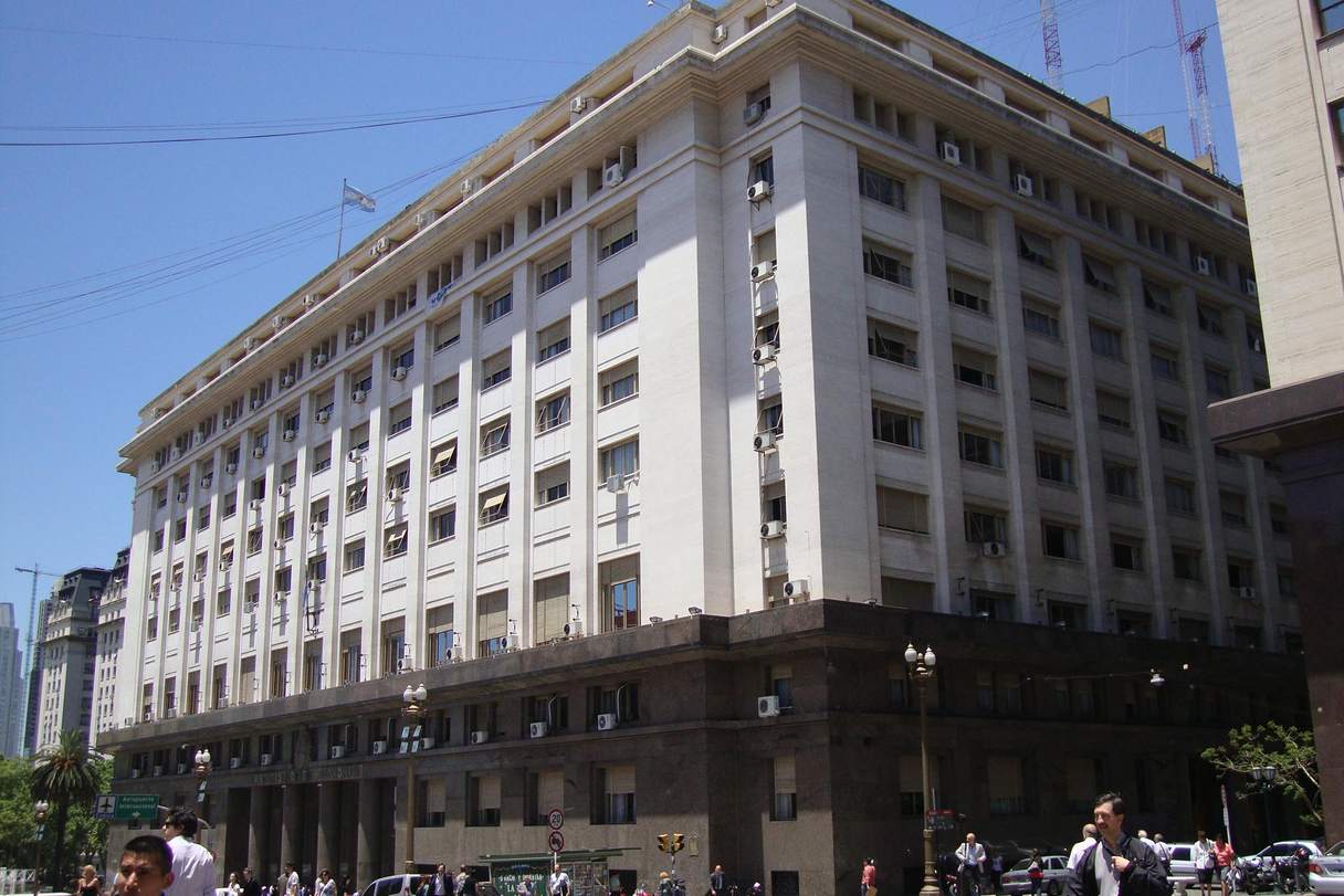 Edificio público de nueve plantas en una esquina, con basamento de piedra oscura, fachada clara de pilastras verticales y una bandera sobre la terraza; peatones caminan por la vereda bajo un cielo despejado.